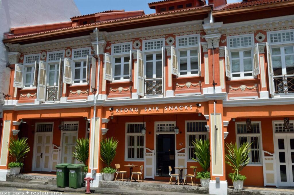 Exterior street view of Keong Saik Snacks, featuring a vibrant orange heritage shophouse building with white shutters and outdoor cafe seating.