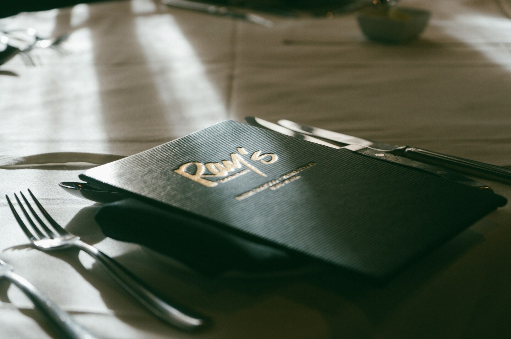 A softly lit restaurant table featuring a dark menu with elegant golden text. Surrounding the menu are polished silverware on a white tablecloth, creating a refined atmosphere.
