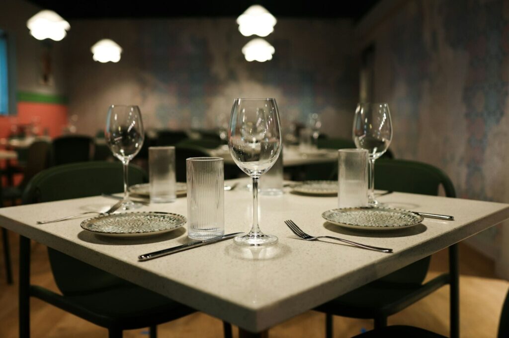 A modern restaurant table set for two with wine glasses, patterned plates, and silverware. Soft lighting creates a cozy, inviting atmosphere.
