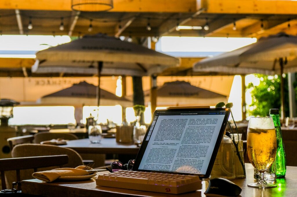 A tablet displays text on a restaurant patio table, accompanied by a keyboard, mouse, and drink. Sunlit outdoor space with a relaxed, peaceful atmosphere.