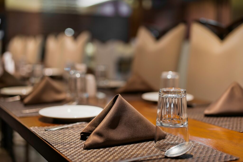 Elegant restaurant setting with neatly folded brown napkins, clear glasses, and white plates on a wooden table, creating a warm, inviting atmosphere.