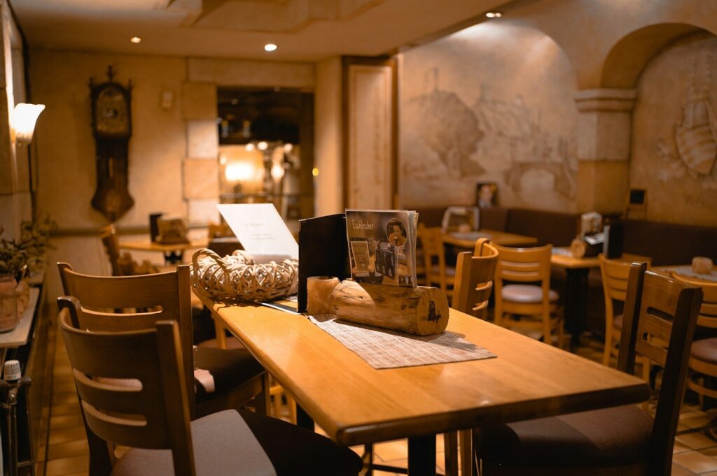 Cozy restaurant interior with warm lighting, featuring wooden tables and chairs. A vintage clock and mural adorn the walls, creating a rustic ambiance.