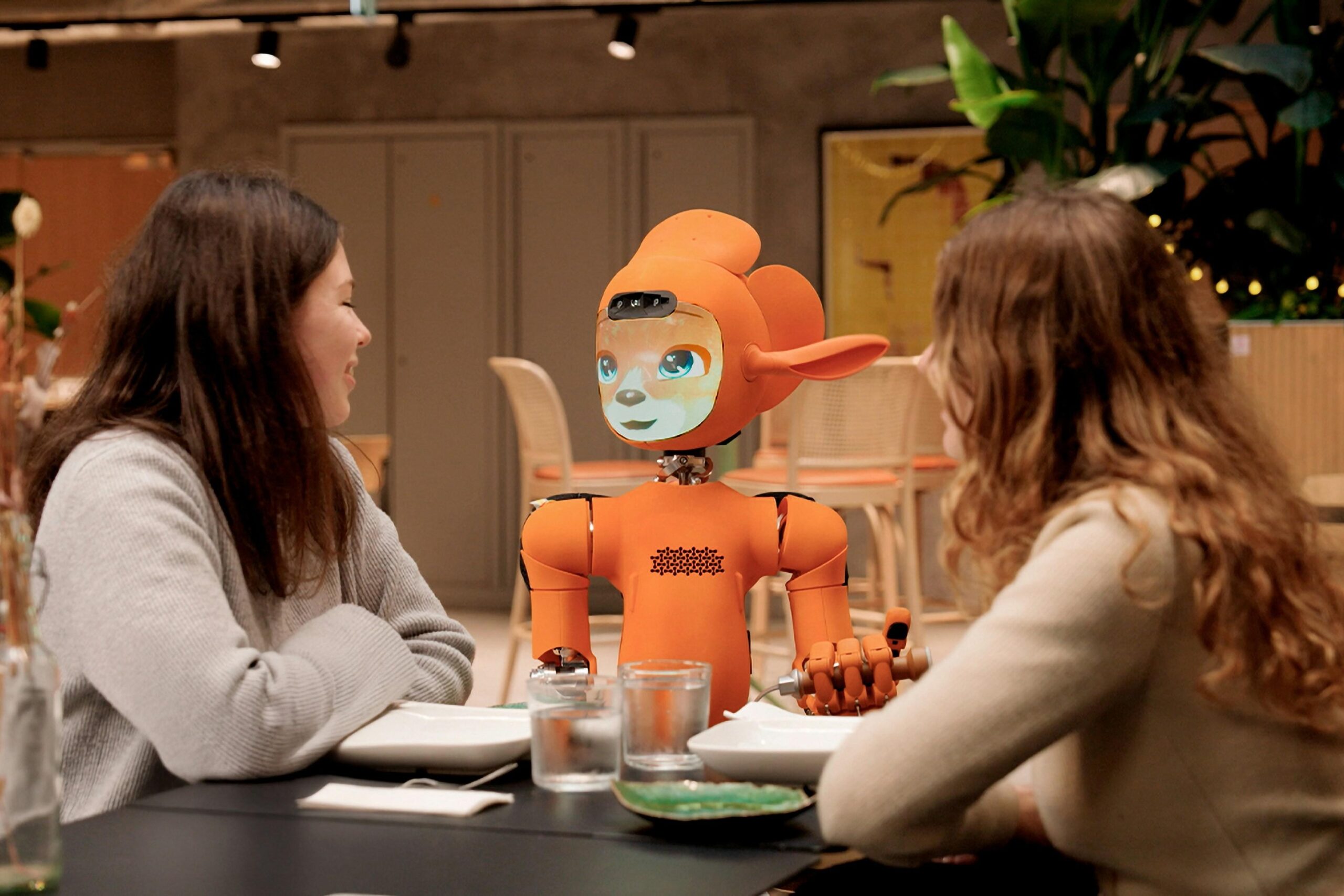 Two smiling women are seated at a table in a modern dining setting, engaging with a bright orange humanoid robot. The robot, featuring an expressive animated face and articulated hands, appears to be participating in the conversation as a friendly companion.