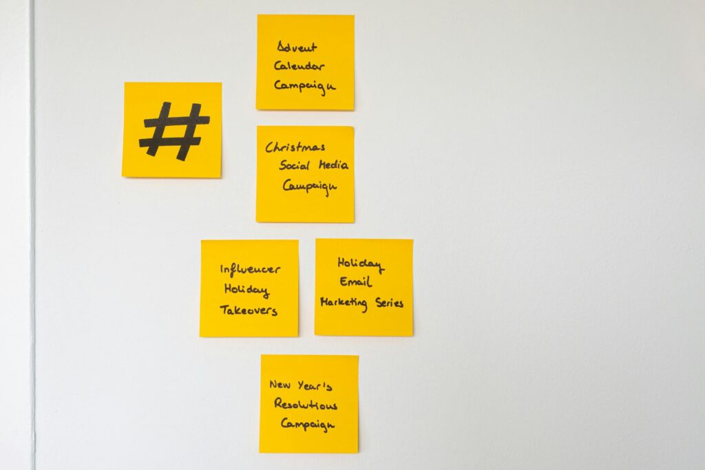 A collection of yellow sticky notes is arranged on a white wall, outlining various holiday-themed marketing strategies such as an "Advent Calendar Campaign" and "Influencer Holiday Takeovers." To the left of the detailed plans, a single note features a large, handwritten hashtag symbol, suggesting a focus on social media content planning.