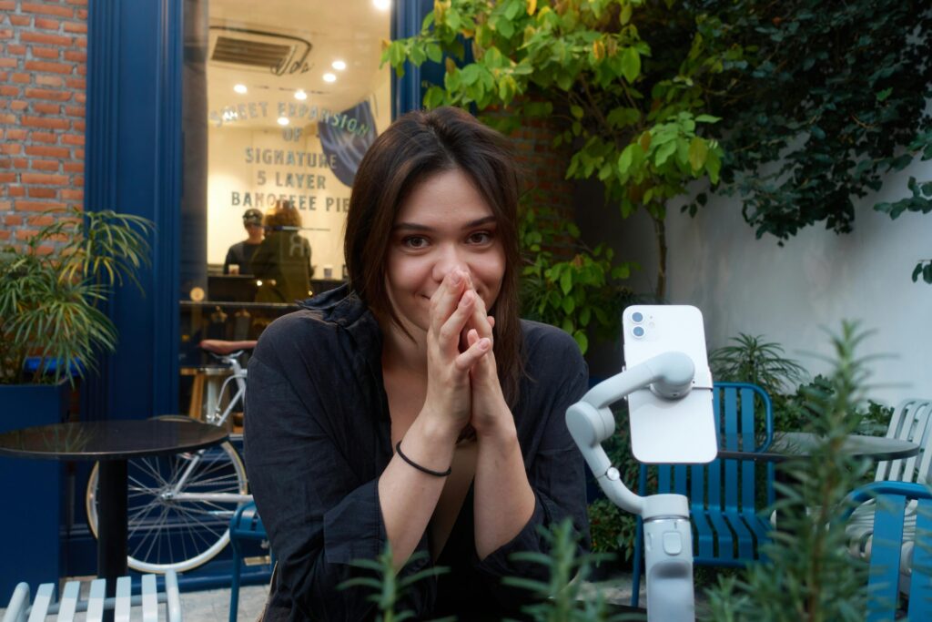 A smiling young woman sits at an outdoor table with her hands clasped, posing in front of a smartphone mounted on a gray gimbal stabilizer. The background features a brick building with a blue framed window advertising banoffee pie, surrounded by lush greenery and patio furniture.