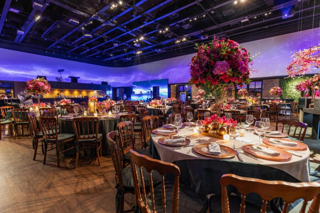 This image captures a lavishly decorated event venue filled with round banquet tables that feature tall, vibrant floral centerpieces and elegant place settings.