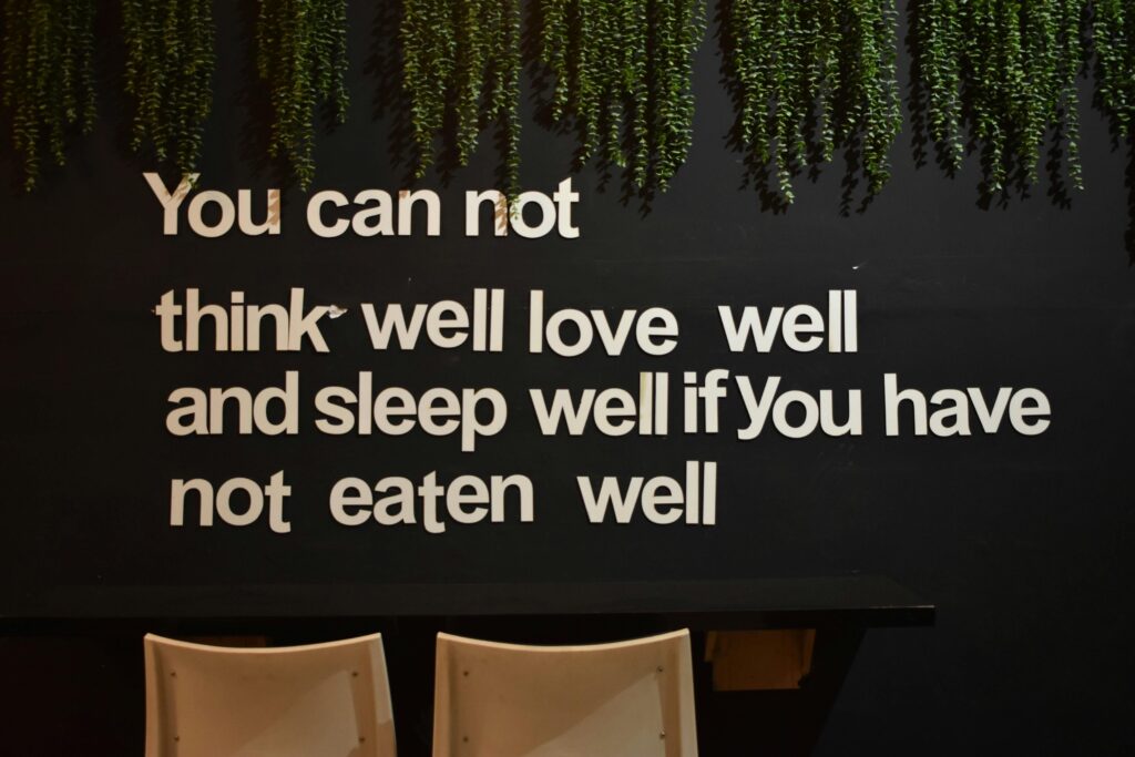 This image features a black wall displaying the quote "You can not think well love well and sleep well if You have not eaten well" in white cutout letters, accented by trailing green plants along the top edge.