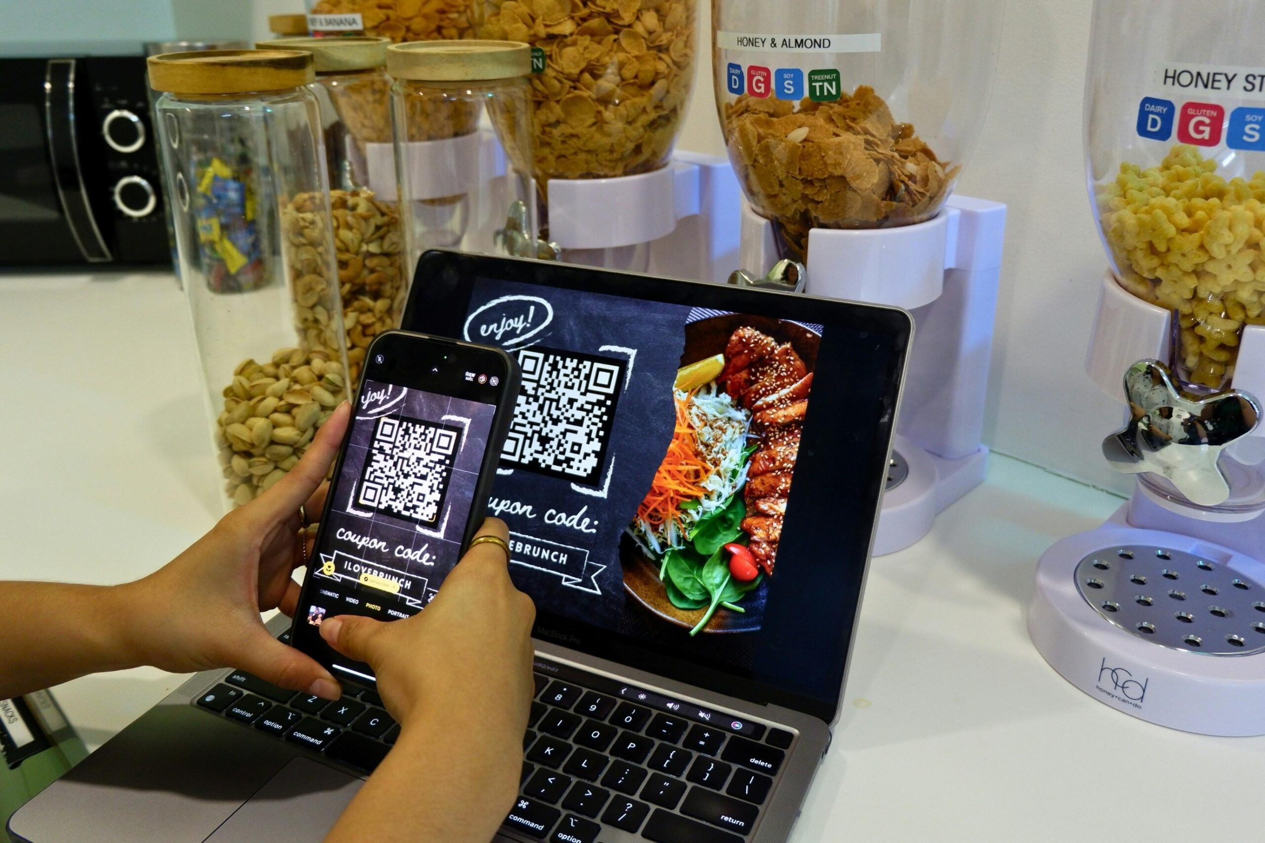 A person uses a smartphone to scan a QR code for a food coupon displayed on an open laptop screen. The setting appears to be a kitchen or breakfast station, featuring a row of dispensers filled with various cereals and nuts in the background.