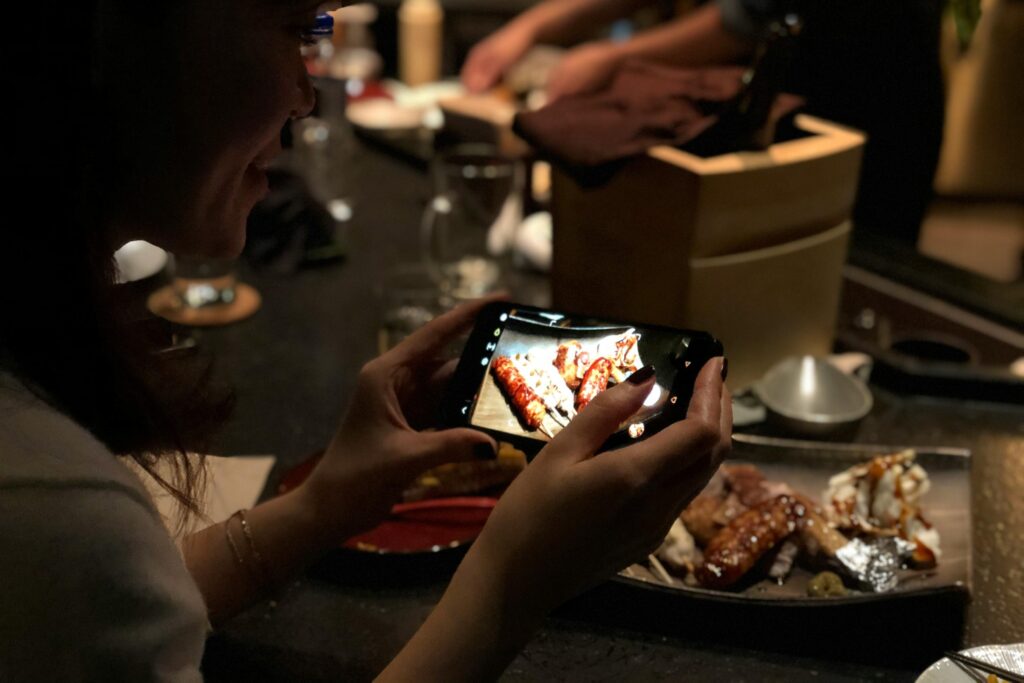 A smiling woman holds a smartphone horizontally to take a picture of a plate of grilled skewers at a dimly lit restaurant counter. The bright phone screen displays the food in focus, contrasting with the warm, dark ambiance of the dining environment.