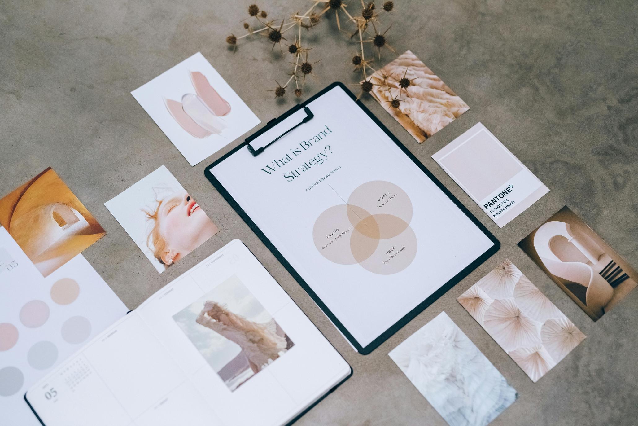 This flat-lay image displays a creative workspace centered around a clipboard titled "What is Brand Strategy?" featuring a Venn diagram. Surrounding the document is a curated arrangement of aesthetic photographs, Pantone color swatches, and a planner, all showcasing a soft beige and peach color palette on a textured gray background.