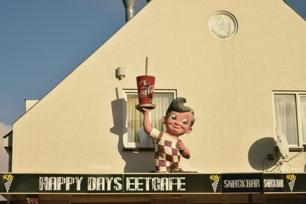 The exterior of the "Happy Days Eetcafe" features a large, retro-style statue of a smiling boy in checkered overalls holding up a giant Coca-Cola cup. Set against a beige brick wall under a clear blue sky, the mascot casts a distinct shadow above the snack bar's black and white signage.