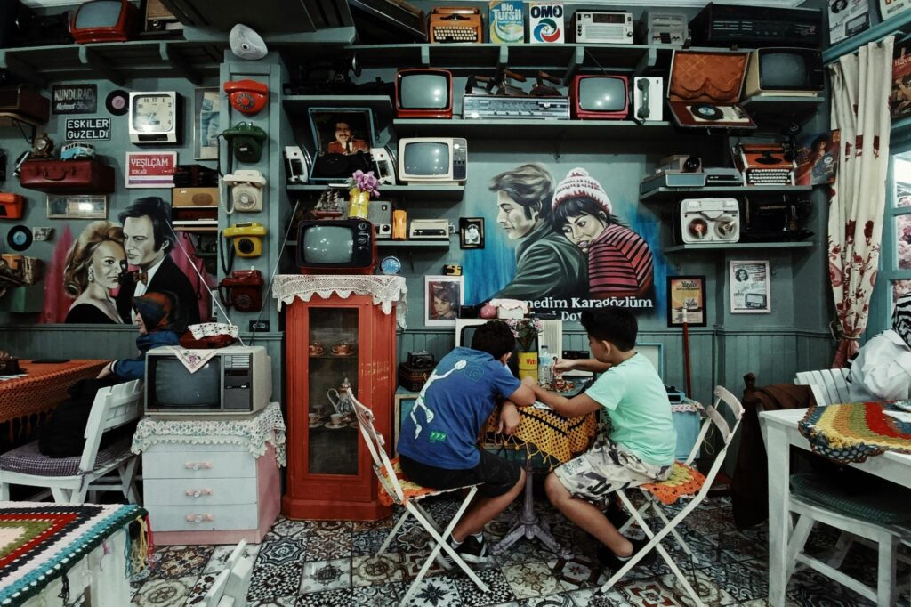 This eclectic cafe interior is densely packed with nostalgia, featuring teal walls lined with shelves holding vintage televisions, rotary telephones, radios, and classic movie posters. In the foreground, two young boys sit at a small table draped with a yellow crocheted cloth, dining amidst the retro decor and intricate patterned tile flooring.