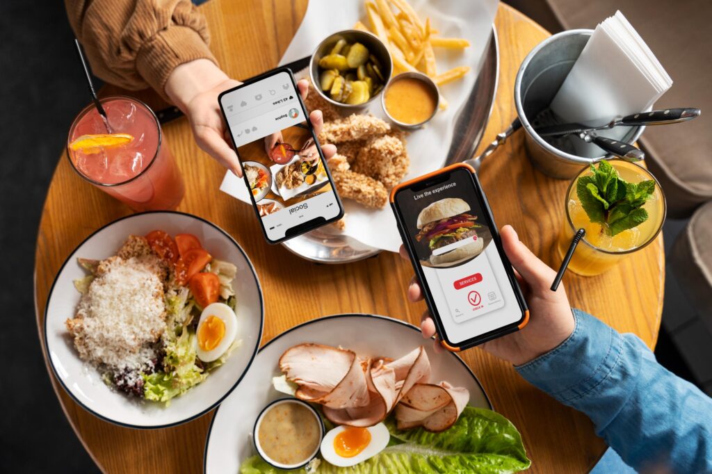 This overhead shot captures two diners holding smartphones over a round wooden table, seemingly documenting their meal or interacting with food-related apps. The table is abundantly filled with appetizing dishes, including fried chicken with fries, fresh salads topped with boiled eggs, and colorful cocktails.