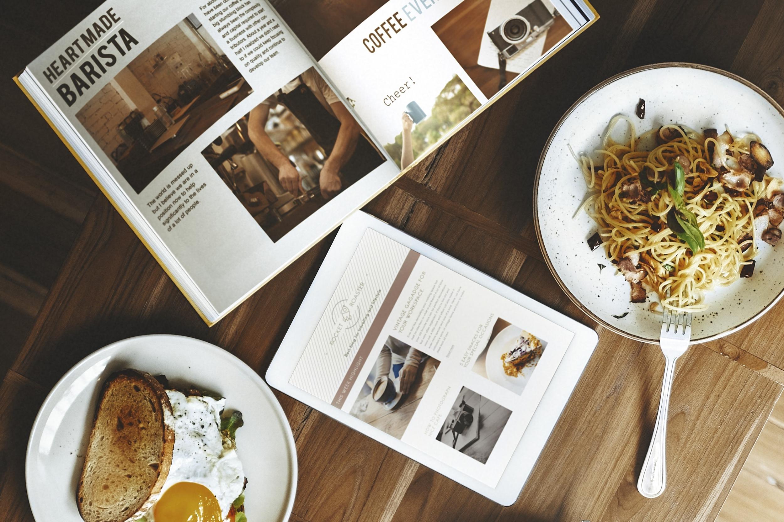 This overhead shot depicts a cozy brunch setting on a wooden table, featuring a plate of pasta and a dish with toast and a fried egg. An open magazine and a tablet displaying lifestyle content lie centrally between the meals, suggesting a relaxed atmosphere for dining and reading.