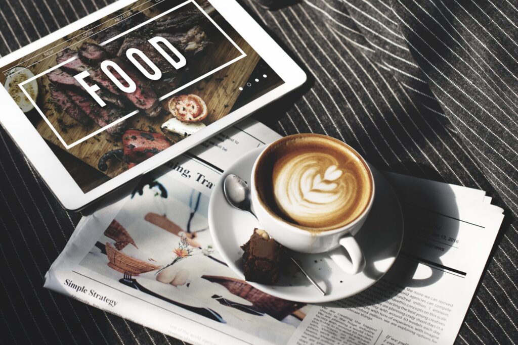 A digital tablet displaying the word "FOOD" alongside a steak image rests on a striped surface next to a cup of coffee with latte art and a small brownie. A newspaper is positioned beneath the coffee cup, creating a cozy scene that blends traditional reading with digital browsing during a relaxing break.