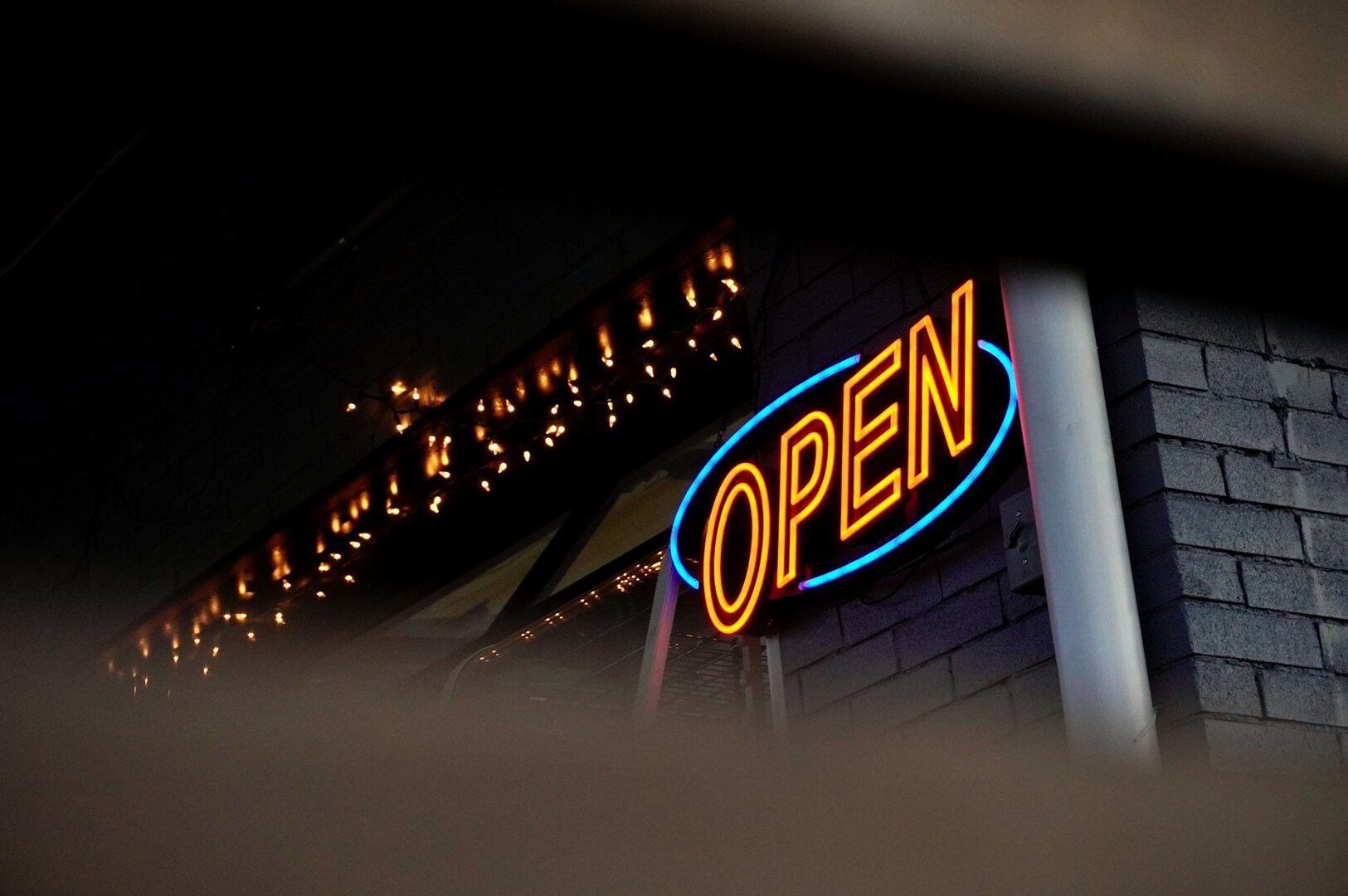 Neon "OPEN" sign glowing in orange and blue against a dark brick wall, with soft twinkling lights reflected in a nearby window, creating a welcoming ambiance.