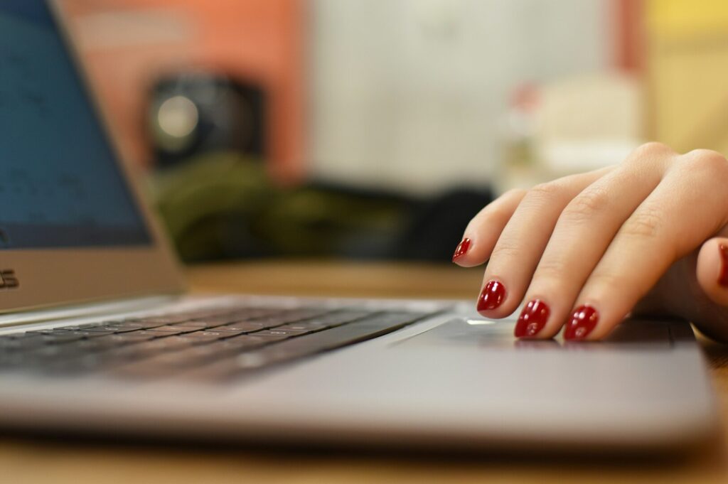 A hand with red nail polish types on a laptop keyboard. The background is softly blurred, conveying a focused, professional atmosphere.