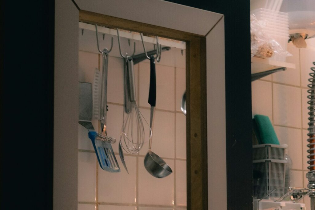 Close-up of hanging kitchen utensils including a whisk, spatula, and ladle against white tiled wall. The scene feels cozy and functional.