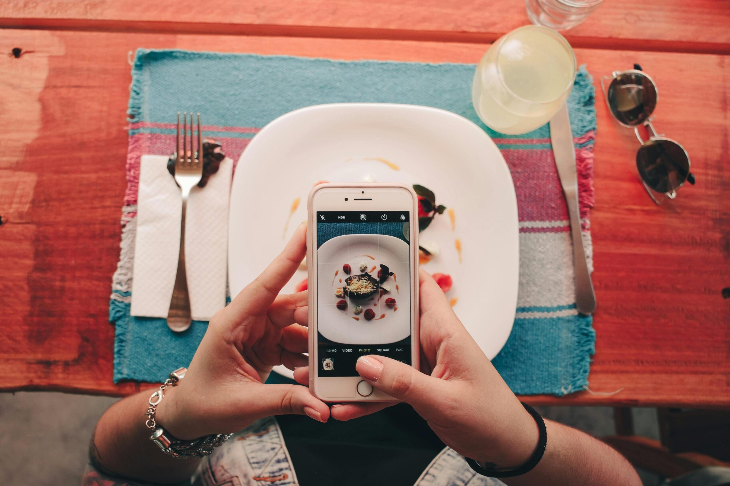 Captured from an overhead perspective, a person uses a smartphone to take a picture of an artistically plated meal sitting on a blue woven placemat. The scene is set on a rustic red wooden table and includes dining essentials like silverware, a napkin, sunglasses, and a glass of a yellow beverage.