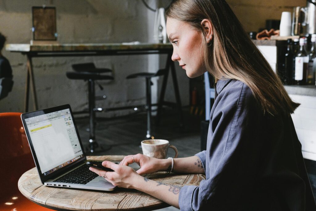 A focused woman sits at a small, round wooden table in a cafe, multitasking between an open laptop and the smartphone held in her hands. She wears a grey shirt with visible arm tattoos and works beside a ceramic mug, while the background features bar stools and counter items typical of a coffee shop setting.