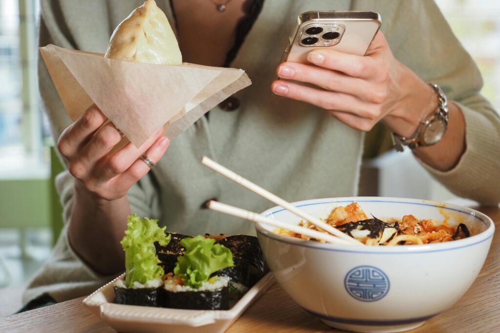 A person seated at a wooden table holds a steamed bun in one hand while simultaneously checking a smartphone held in the other. In the foreground, a spread of Asian cuisine is displayed, featuring a bowl of noodle soup with chopsticks resting on top and a side of sushi rolls.