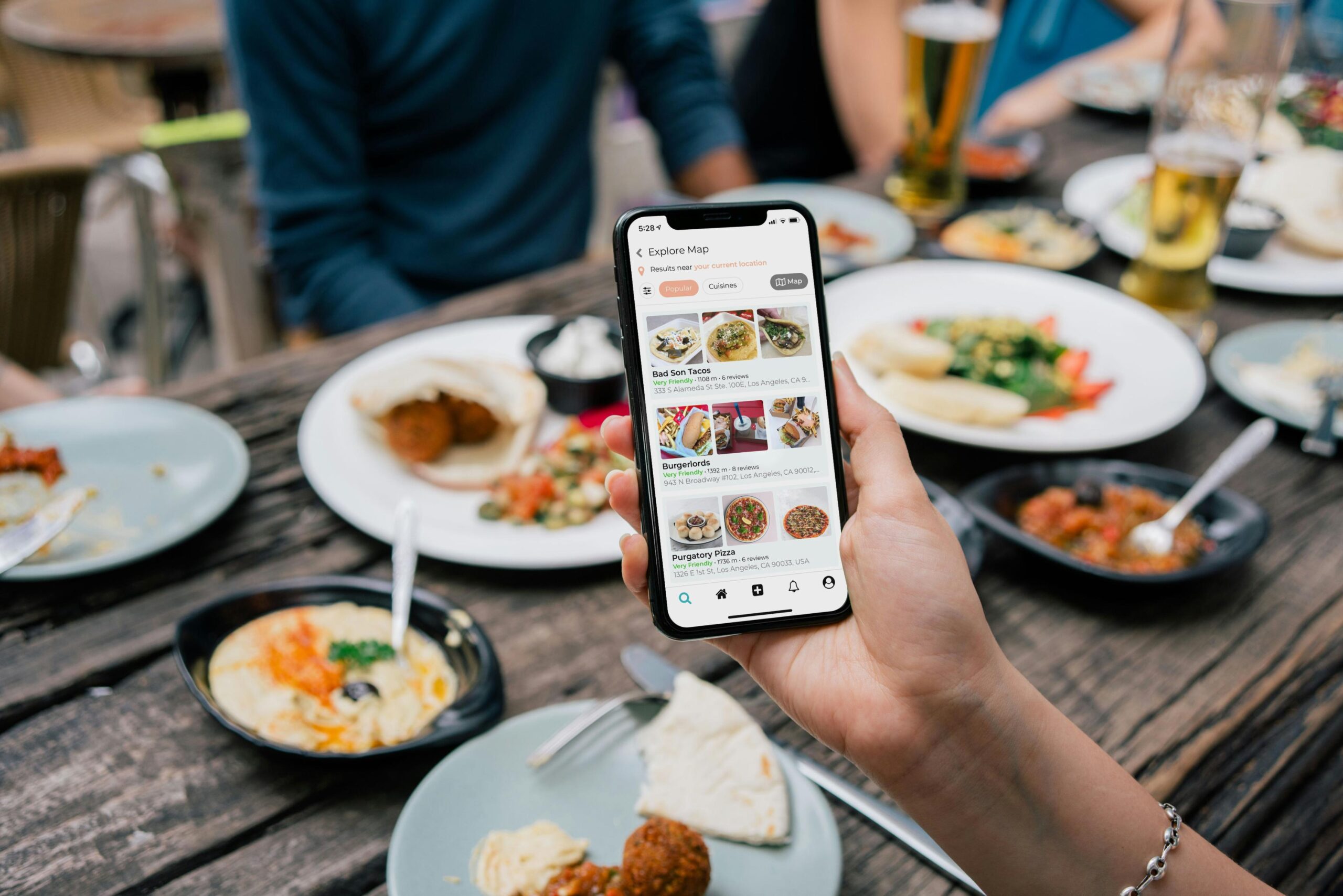 A close-up view captures a hand holding a smartphone that displays a food discovery app with various restaurant listings and photos. The background reveals a lively dining scene at a wooden table, cluttered with plates of food, drinks, and other diners enjoying a meal.