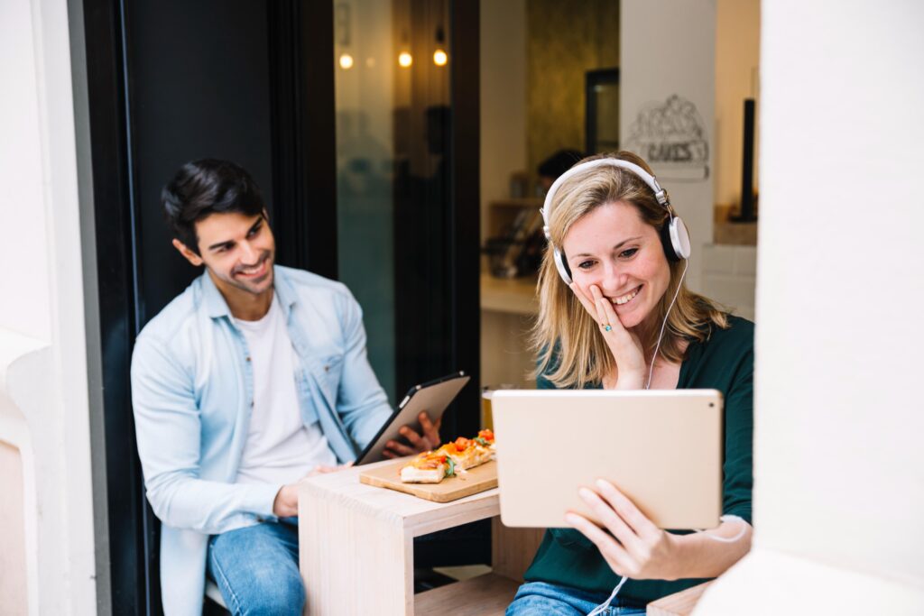 A cheerful woman wearing headphones laughs while looking at a tablet screen in the foreground, while a man seated behind her holds his own device and smiles. They appear to be relaxing at a cafe table with a wooden board of appetizers placed between them.