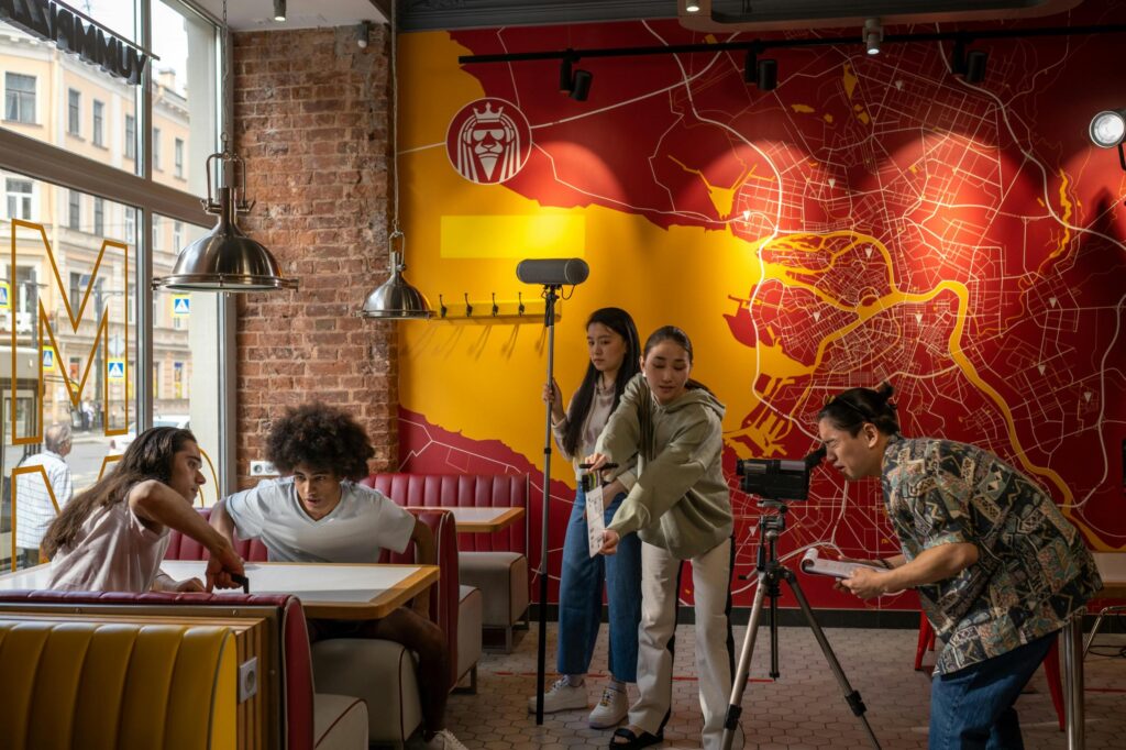 A film crew is actively shooting a scene inside a diner that features a large red and yellow map mural on the wall, with two actors seated in a booth. The production team surrounds the actors, managing essential equipment including a boom microphone, a clapperboard, and a camera mounted on a tripod.