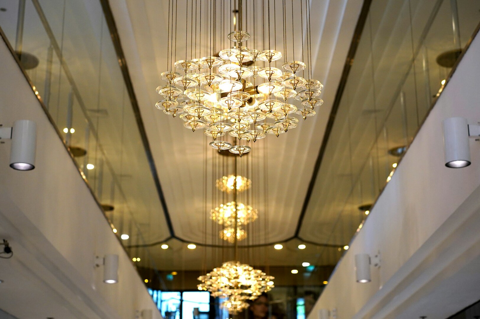Elegant chandeliers with crystal glass details hang in a mirrored corridor, reflecting warm light. The setting is luxurious and sophisticated.