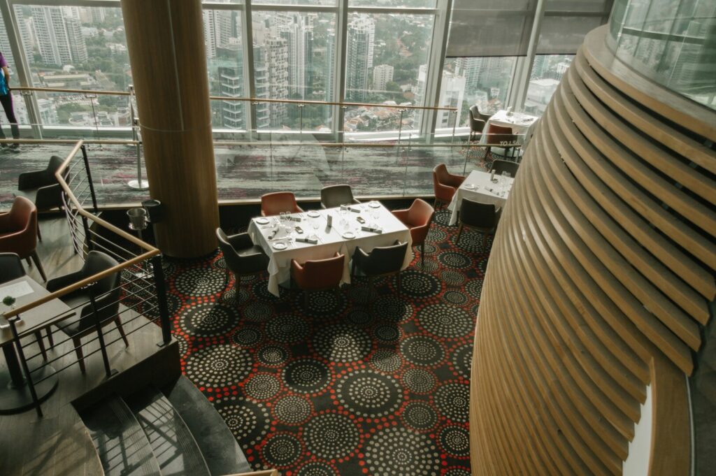 A high-rise restaurant with a modern design, featuring circular patterned carpet, elegantly set tables, and a panoramic city view through large windows.