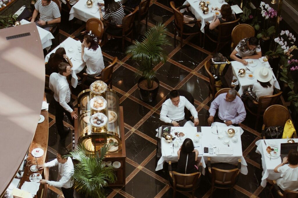 Bird's-eye view of a stylish café with people dining at tables draped in white cloths. Lush plants are interspersed, creating a relaxed, lively ambiance.