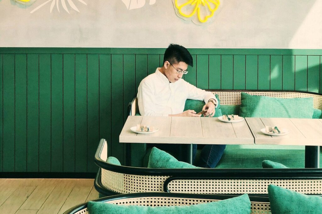 A man in a white shirt sits alone at a green-themed cafe. He checks his phone at a table set with plates. The mood is calm and focused.