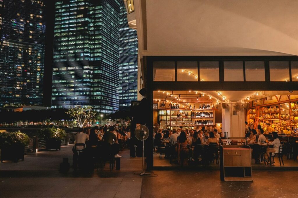 Outdoor evening scene with people dining at a warmly lit restaurant beside modern skyscrapers. Ambiance is lively and urban against the city backdrop.