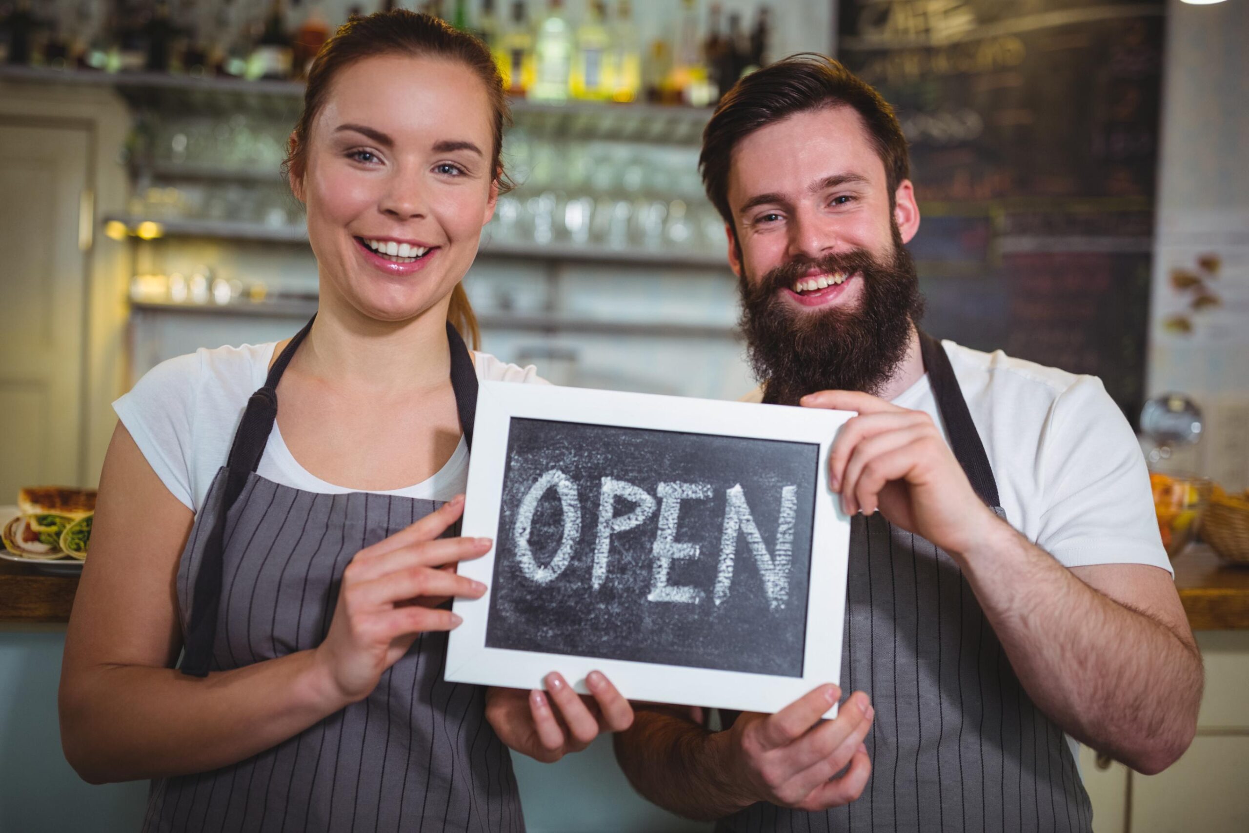 A smiling woman and a bearded man, both wearing matching grey aprons, stand together inside a cafe.