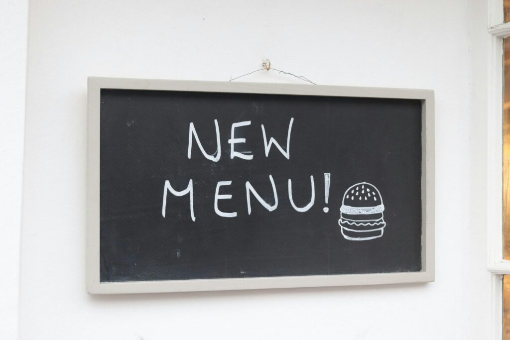 A rectangular chalkboard sign with a light frame hangs against a clean white wall, displaying the handwritten message "NEW MENU!".