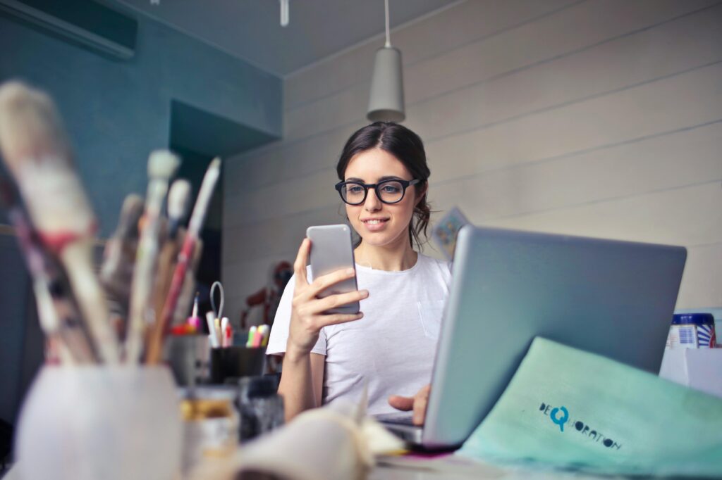 A smiling woman wearing glasses sits at a desk, checking her smartphone while simultaneously keeping one hand on her laptop keyboard.