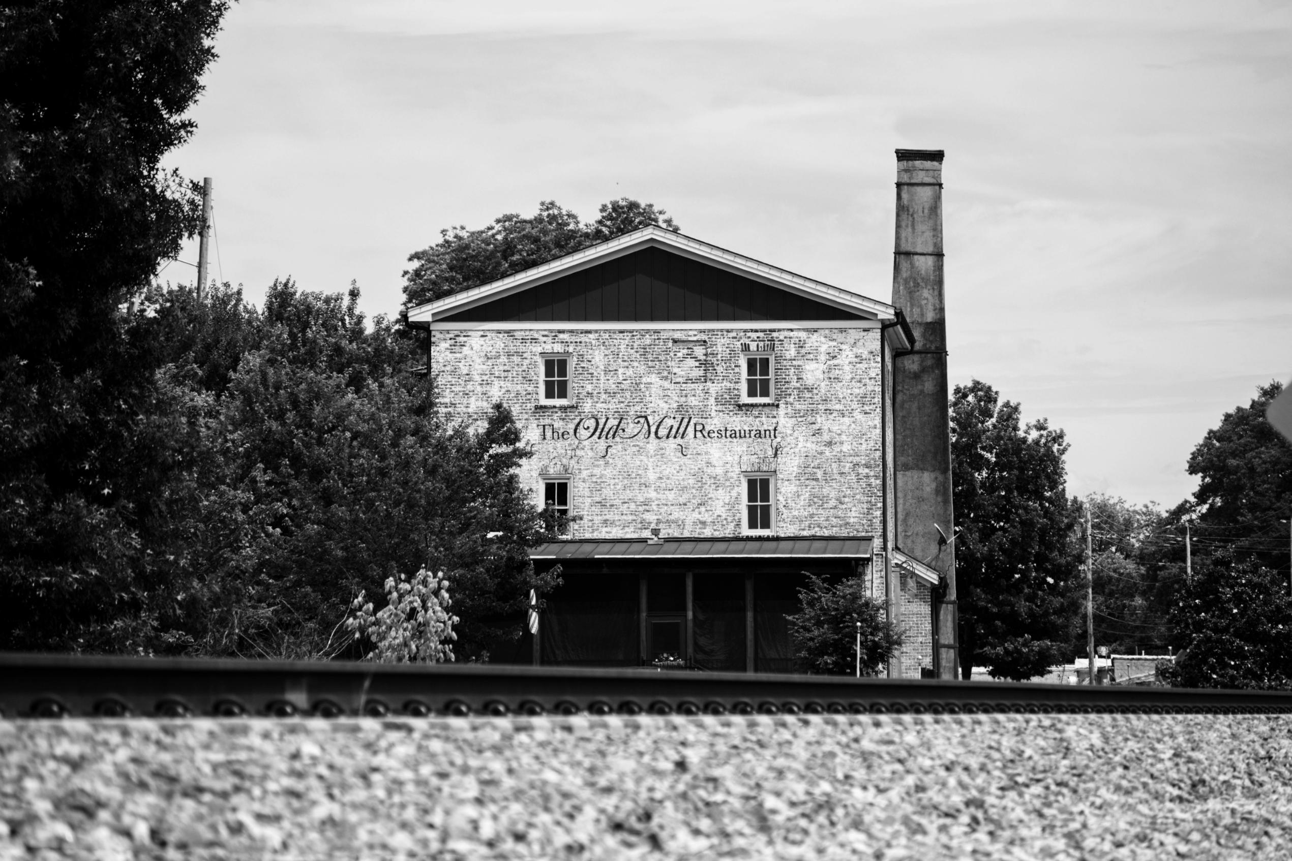This black-and-white photograph captures a weathered brick building identified as "The Old Mill Restaurant," which features a tall chimney stack and is surrounded by dense trees.