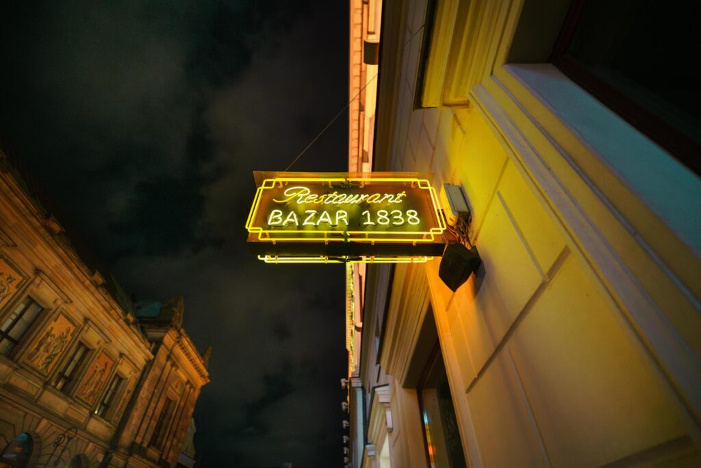 A bright yellow neon sign reading "Restaurant BAZAR 1838" hangs from the side of a building, casting a warm glow on the surrounding architecture.