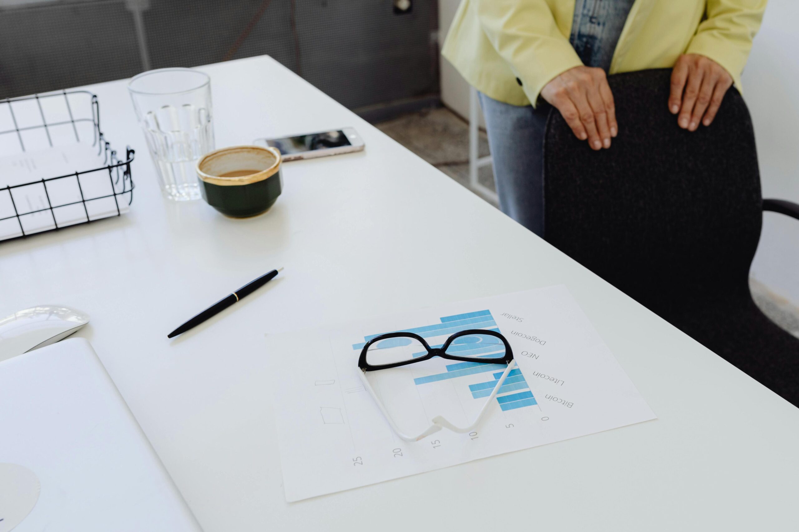 A pair of black-rimmed eyeglasses rests on a printed cryptocurrency bar chart on a white office desk, surrounded by a pen, a coffee cup, and a glass of water.