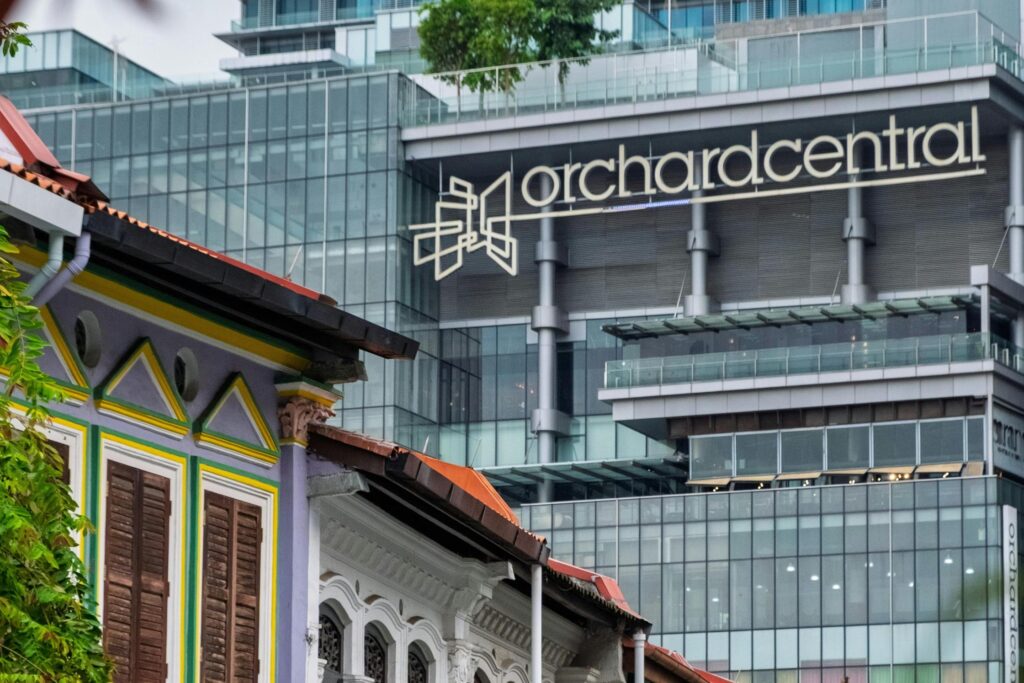 This image displays a striking architectural contrast, featuring the colorful, ornate facade of a traditional shophouse in the foreground set against a towering modern glass structure.