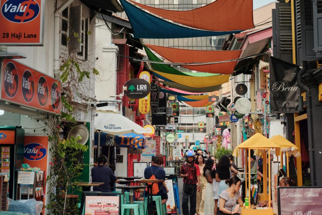 This vibrant, narrow street scene features a bustling crowd of pedestrians and diners amidst a colorful array of shops and restaurants, marked by prominent signage like "Valu$" and "Lira."