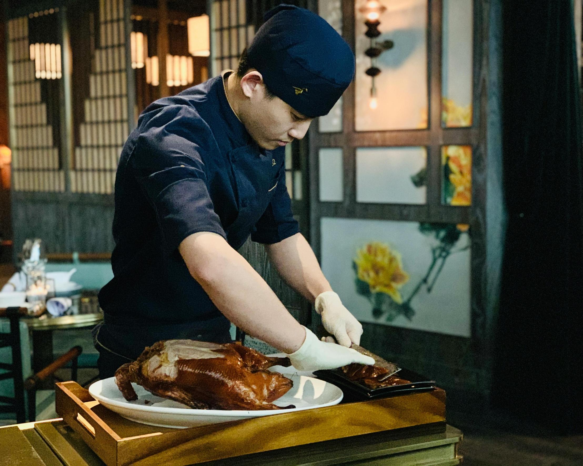 A chef in a dark blue uniform and white gloves meticulously carves a whole roasted duck at a table.