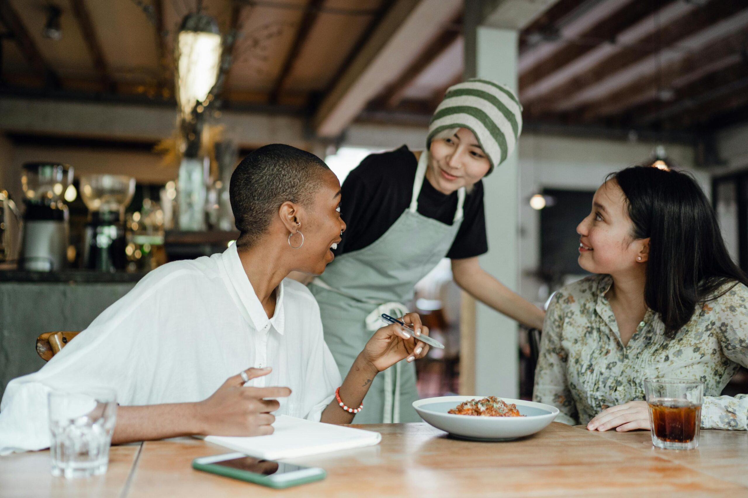 Two women sit at a wooden table in a cafe, engaging in a lively conversation with a server wearing a striped beanie and apron.