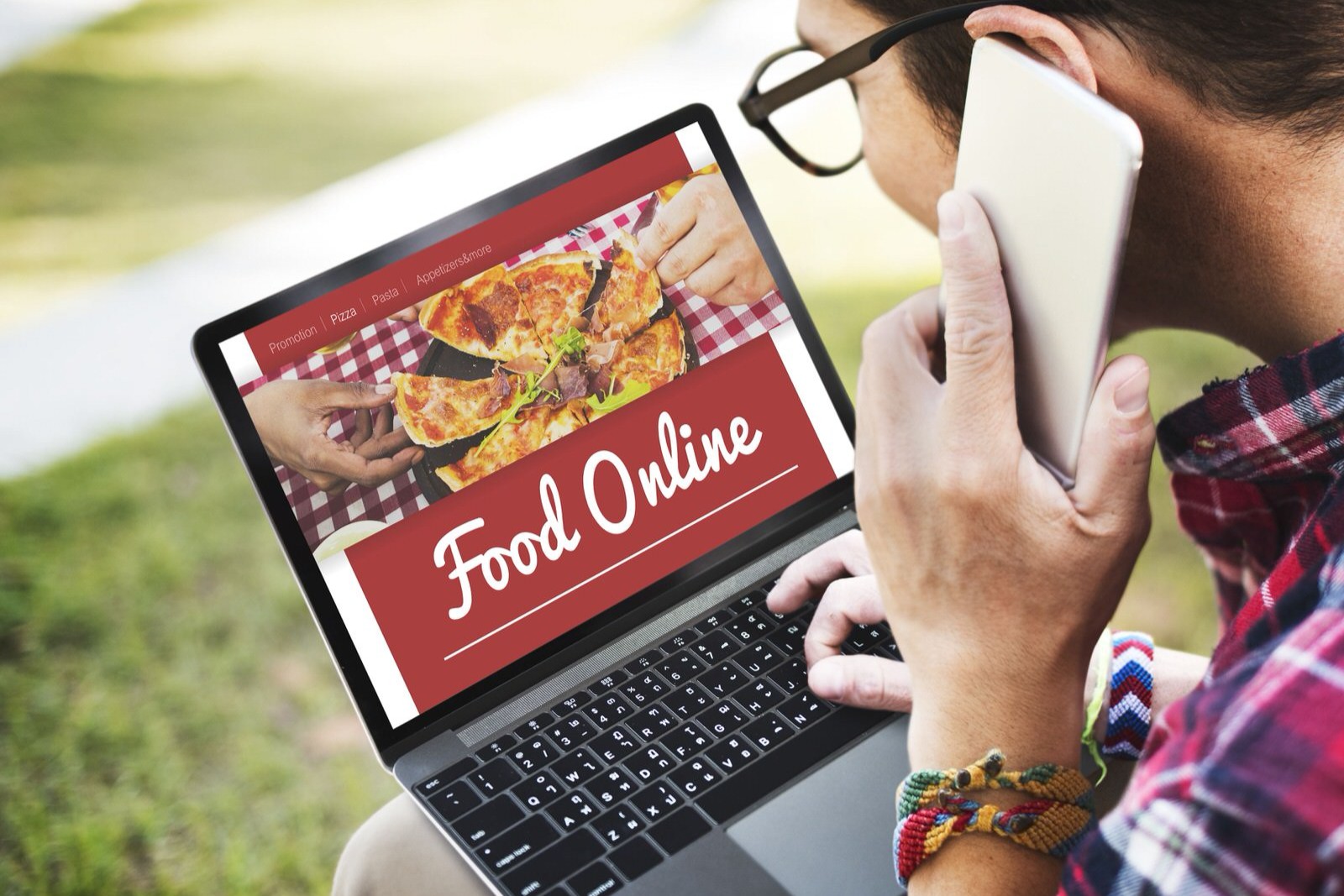 A person in a red plaid shirt is sitting outdoors, holding a phone to their ear while looking at a laptop. The laptop screen displays a "Food Online" website featuring a large image of a pizza.