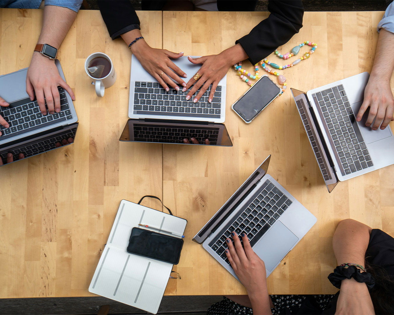 Several hands rest beside open laptops on a shared table, showing a group actively collaborating and exchanging ideas during a project discussion.