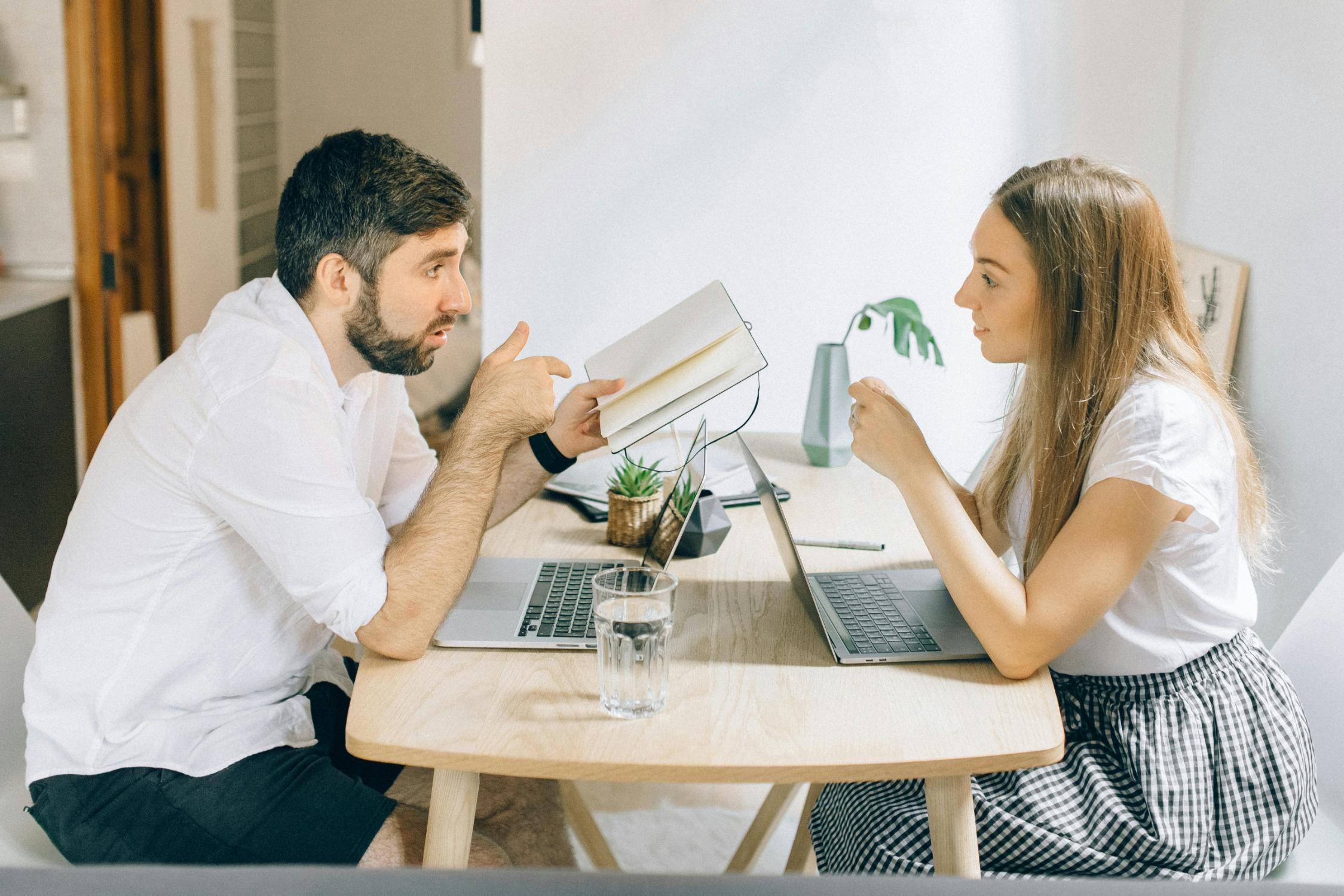 A man and a woman sit across from each other at a light wood table, both working on laptops in a bright, minimalist room.