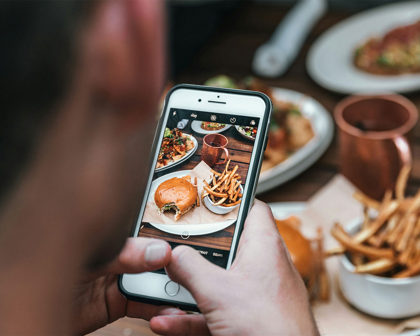 A man holding his smartphone above a beautifully plated dish, taking a photo to capture the food’s presentation. The table is set elegantly, with soft ambient lighting highlighting the dish.