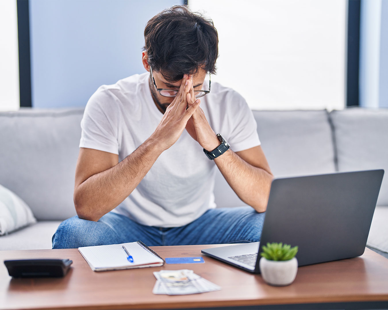 A man sits in front of his laptop with his hands on his head, showing a stressed and overwhelmed expression while working indoors.