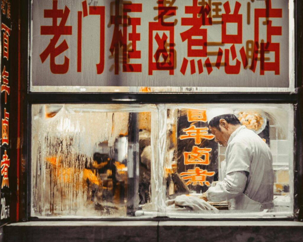 A man in white kitchen attire works behind a steamed-up window of a restaurant adorned with large red and orange Chinese characters.