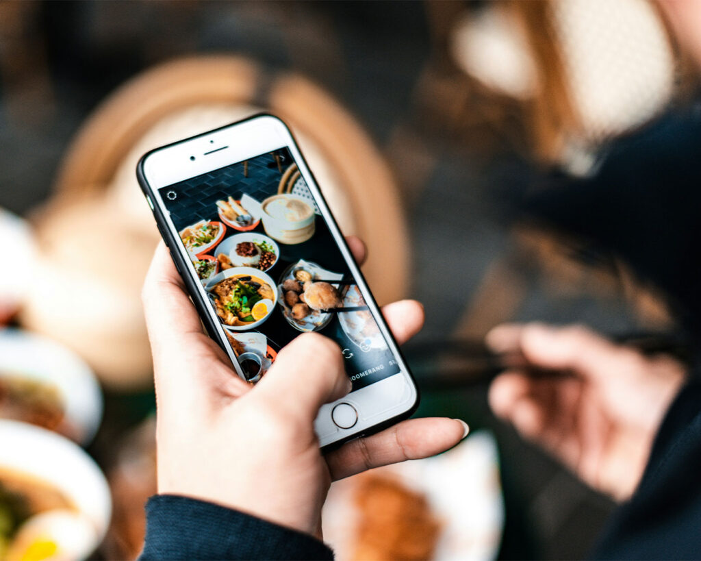 A hand holding a smartphone capturing a photo of a plated dish in a restaurant, showing food content creation or social sharing.